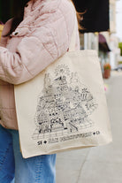 A person wears a pink jacket and jeans, clutching the Cat House Tote adorned with a whimsical San Francisco illustration reminiscent of Ferris Plock's unique style.