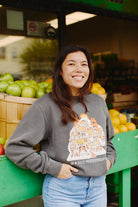 A person in a Cat House Crewneck smiles in front of a fruit stand filled with baskets of apples and oranges.