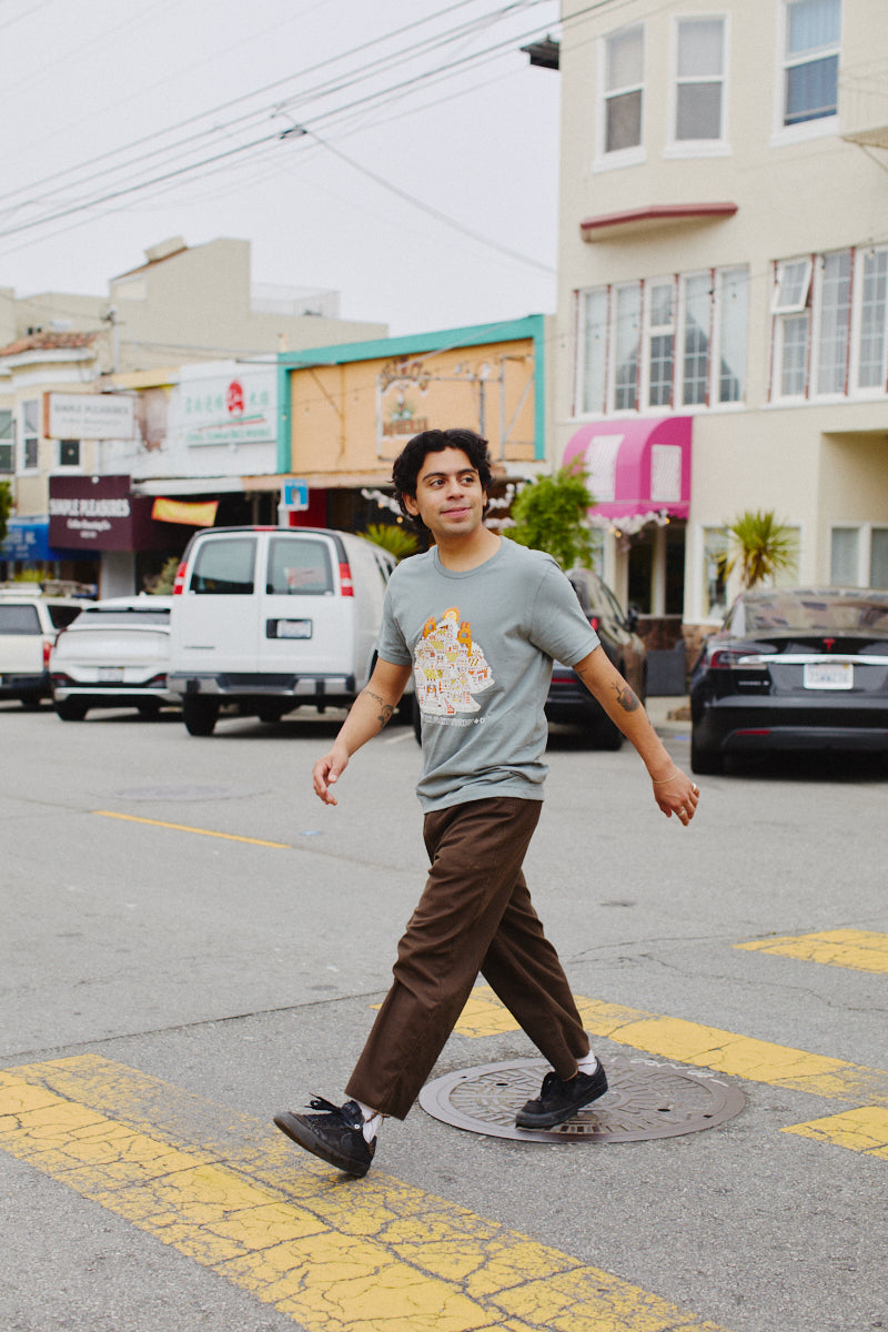 A person crosses a bustling urban street wearing the unisex Cat House Tee, made from soft cotton, as shops and cars animate the background.