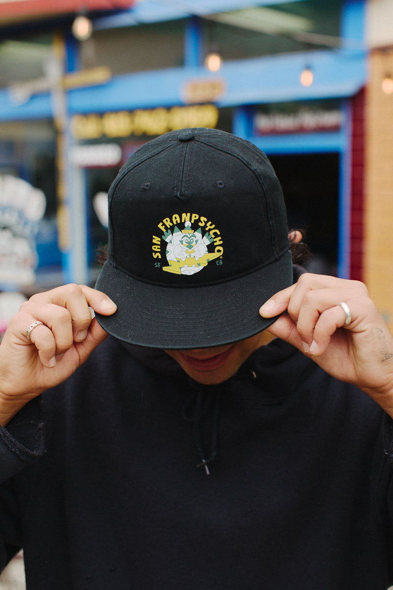 A person is wearing the black Lightning Cat Hat, featuring a San Francisco design, and is adjusting the brim outdoors against a vibrant background reminiscent of Ferris Plock's artwork.