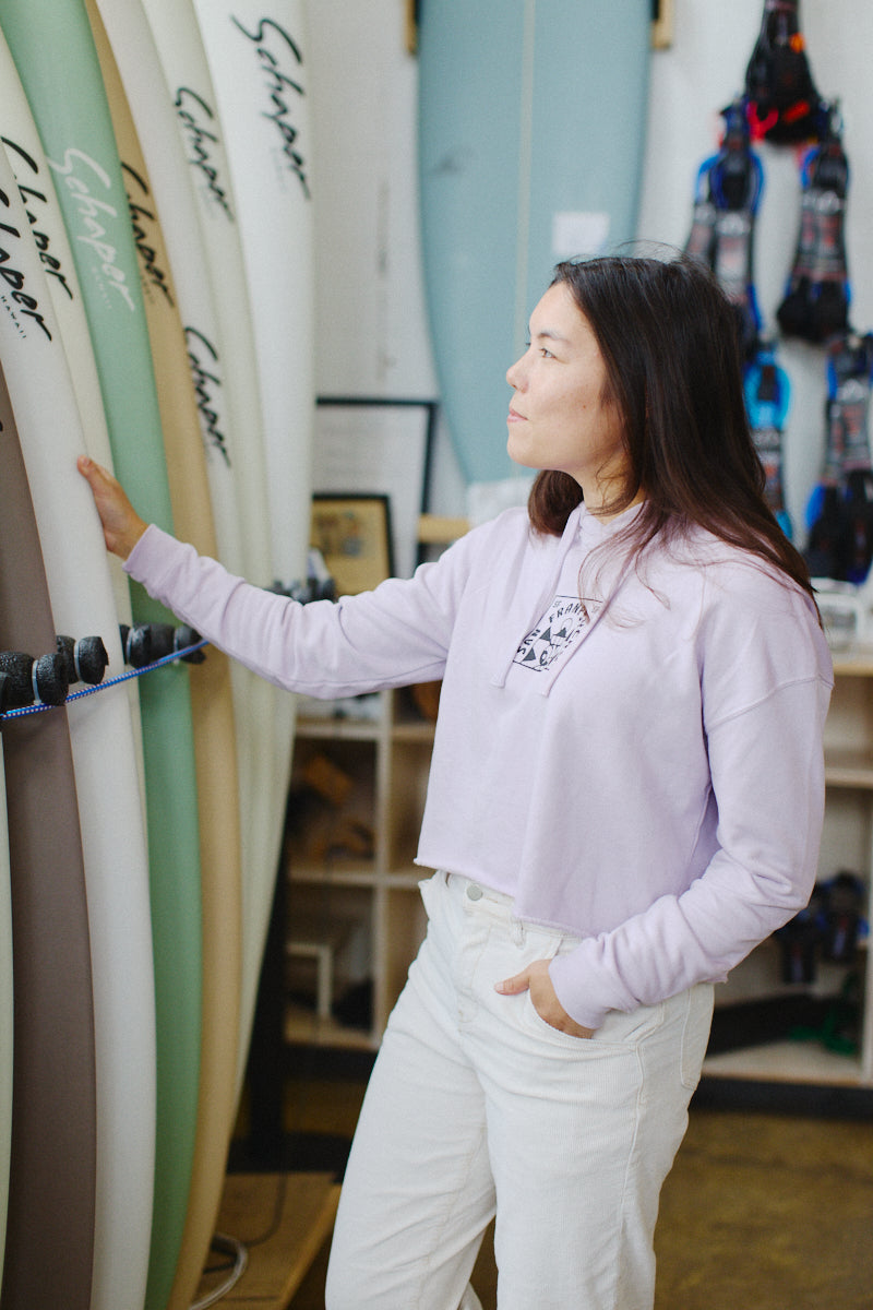 A person in a Lavender Cat Stamp Crop Hoodie is browsing surfboards in a shop.