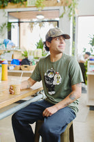 A person sits at a café table, donning a Battle Cat Tee and matching cap while holding an iced coffee. Vibrant Ferris Plock artwork and lush plants surround them, blending nature with creativity.