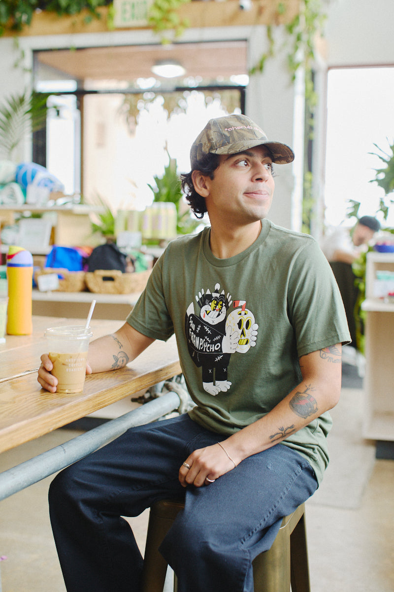 A person sits at a café table, donning a Battle Cat Tee and matching cap while holding an iced coffee. Vibrant Ferris Plock artwork and lush plants surround them, blending nature with creativity.