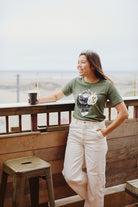 A woman in a Battle Cat Tee smiles while holding a coffee cup on a wooden deck near the beach.