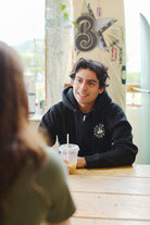 A smiling man in a black Unisex Battle Cat Zip-Up Hoodie, seated at a table with a drink, is surrounded by surfboards.