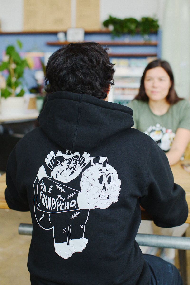 A person wearing a Unisex Battle Cat Zip-Up Hoodie showcasing Ferris Plock artwork sits at a table across from another individual in a comfortable indoor environment.