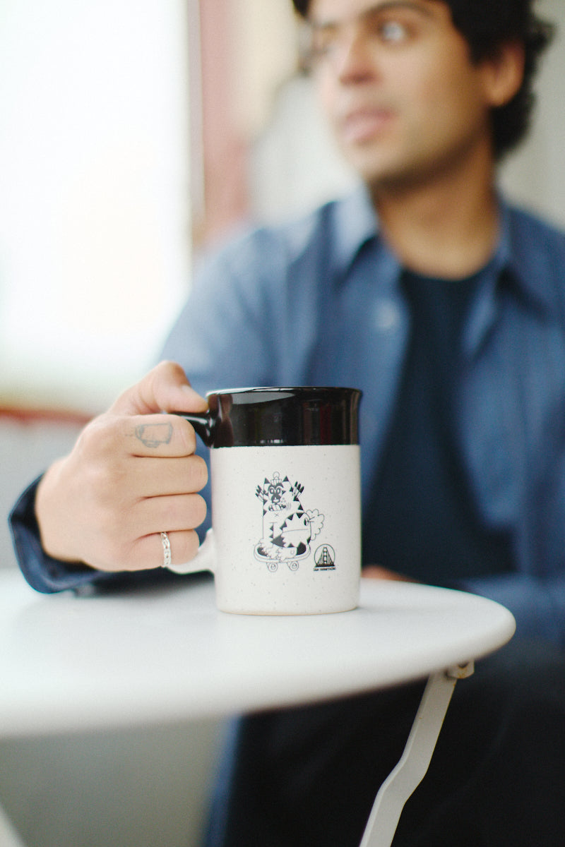 A person sits at a small round table, holding the charming Kitty Poof Mug, which features an intricate black design.