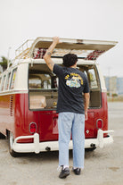 A person is positioned at the open rear door of a red and white vintage van, sporting the Surf Trip Tee, crafted entirely from Airlume cotton. This unisex tee exudes a relaxed vibe that's ideal for any adventure.