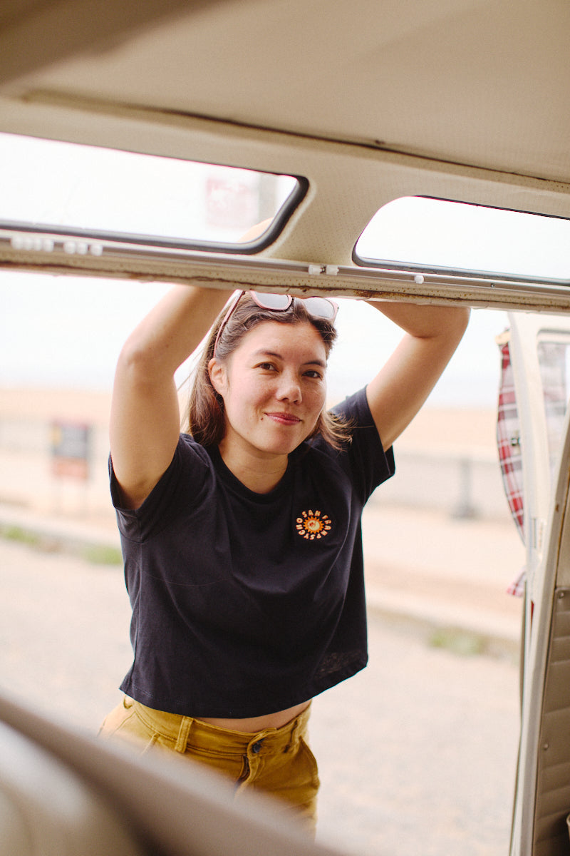 Woman smiling, holding the door frame, stands outdoors near a beach, her Sunny Crop Top adding a touch of elegance that complements her vibrant surroundings.