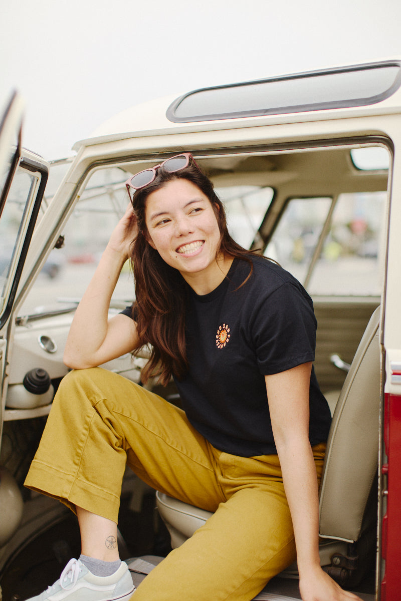 A smiling person sitting in a car doorway, showcasing the Sunny Crop Top in black, paired with mustard pants, exudes a style reminiscent of Ferris Plock's artwork.