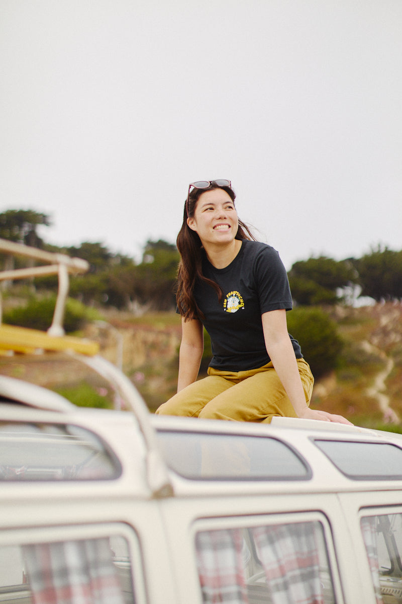 Wearing her Surf Trip Tee made from 100% Airlume cotton, a woman sits smiling on top of a van as the breeze flows through her shirt, with trees and rocky landscape creating an ideal backdrop.