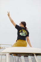 Perched atop a van roof, an individual sports the Surf Trip Tee, crafted from 100% Airlume cotton, while flashing a peace sign with one hand.