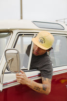 A person leans out of a red and white van window, sporting the Surf Trip Hat in yellow with its unstructured crown. Tattoos adorn their arm, whispering tales of numerous adventures and surf excursions.