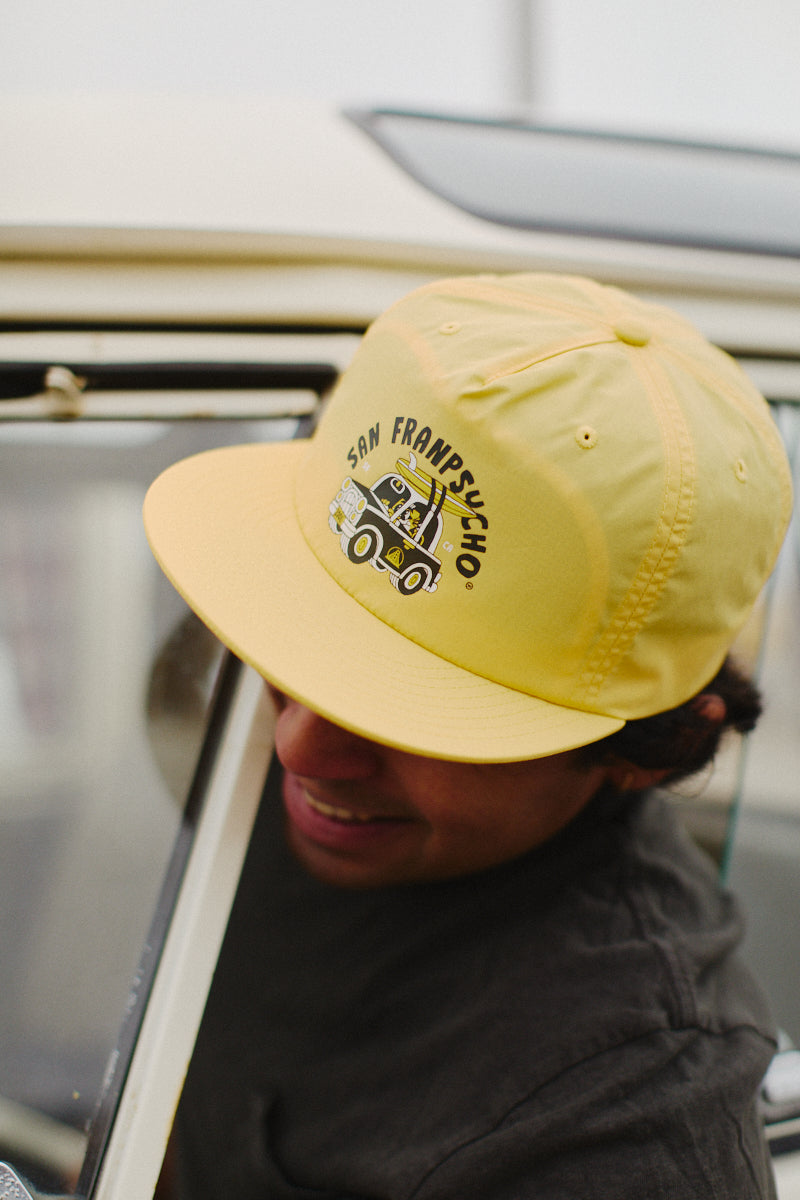 A person wearing a Surf Trip Hat, which features a yellow cap with a playful cartoon design and the San Franpsycho text, gazes out of a car window. Its unstructured crown and flat brim contribute to its relaxed style, making it ideal for soaking up coastal vibes while traveling.