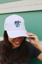 A smiling person is wearing the Kitty Poof Dad Hat, which showcases a white cap adorned with a blue and orange embroidered patch, perfectly complemented by an adjustable back for comfort.