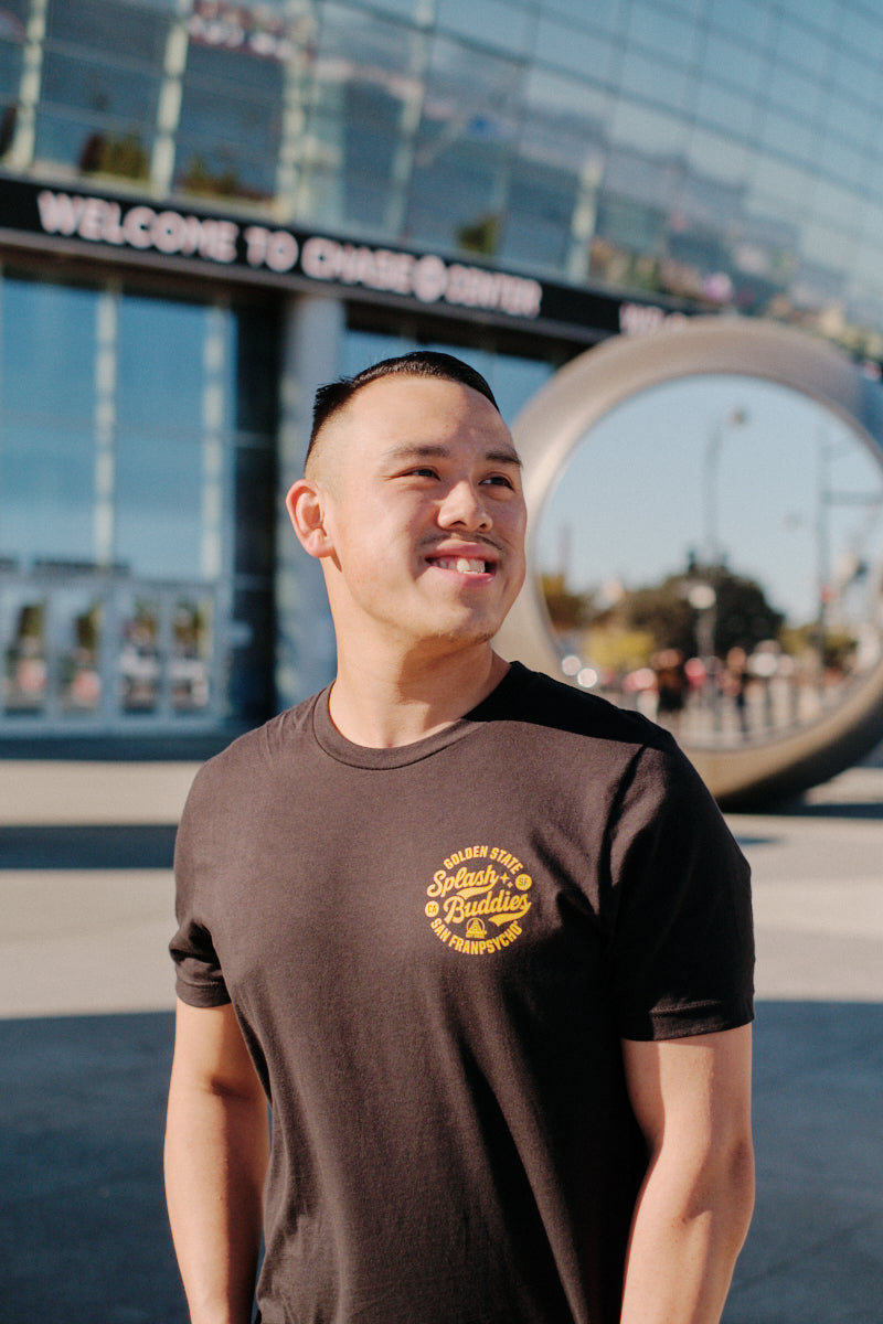 A person smiles while standing in front of a modern building with glass windows, wearing a Splash Buddies Tee. A large metal sculpture and a sign that reads "Welcome to Chase Center," home of the Warriors, are visible in the background.