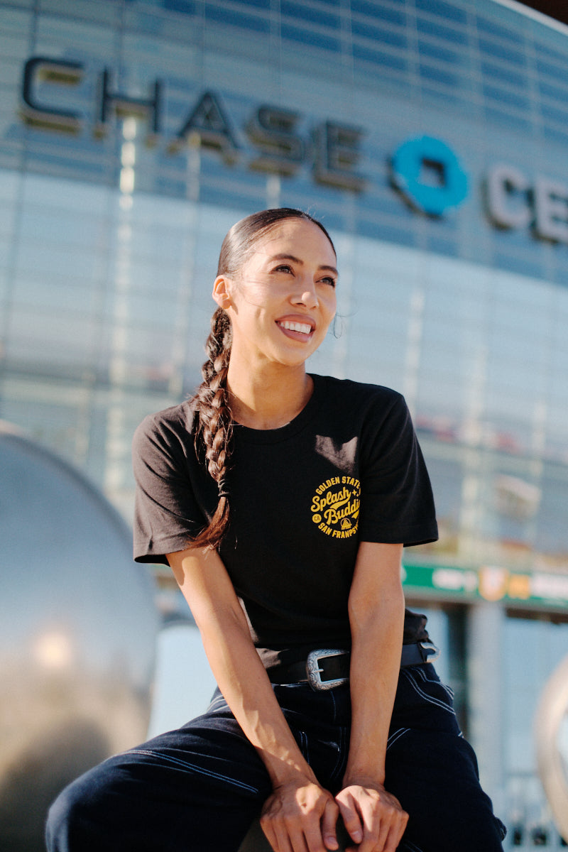 A woman with a braided hairstyle is sitting and smiling in front of the Chase Center, enjoying the sunny day. She's wearing the Splash Buddies Tee, a Warriors unisex shirt featuring bold yellow text—perfect for fans of the team.