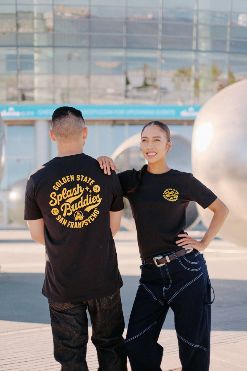 Two individuals wearing black Splash Buddies unisex tees stand outdoors. One smiles while facing forward with hands on hips, and the other turns away. Large metallic spheres and a building serve as the backdrop.