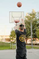 Wearing a Splash Buddies Tee, a person sports a black unisex shirt with yellow lettering as they shoot a basketball towards the hoop on an outdoor court. "Splash Buddies" is prominently displayed on their back, capturing the spirit of true Warriors fans. The scene is perfectly set with trees and a clear sky in the background.