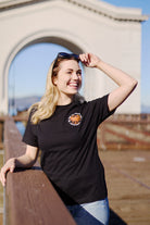 Sporting a Wharf Crab Tee, a smiling woman stands on a pier at Fishermans Wharf, adjusting sunglasses with an archway and hints of the bustling San Francisco scene behind her.