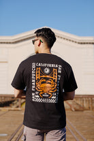 A man stands outdoors on a sunny day, wearing a black Wharf Crab Tee that beautifully captures the essence of Fishermans Wharf in San Francisco.