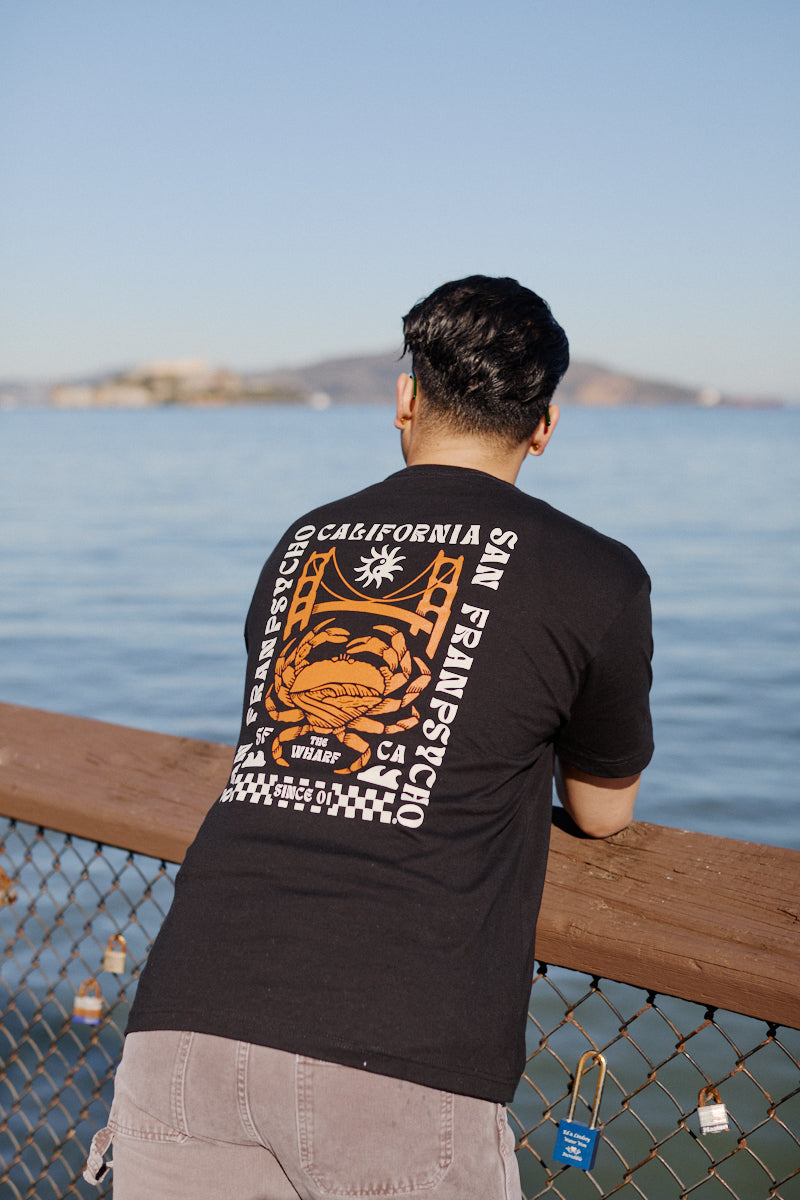 A man in a black Wharf Crab Tee leans on a railing by the water at Fishermans Wharf, with hills in the background.