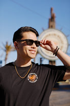 A smiling man in sunglasses dons a Wharf Crab Tee with a gold chain, standing outdoors near San Franciscos Fishermans Wharf, while the blurred background enhances the coastal vibe.