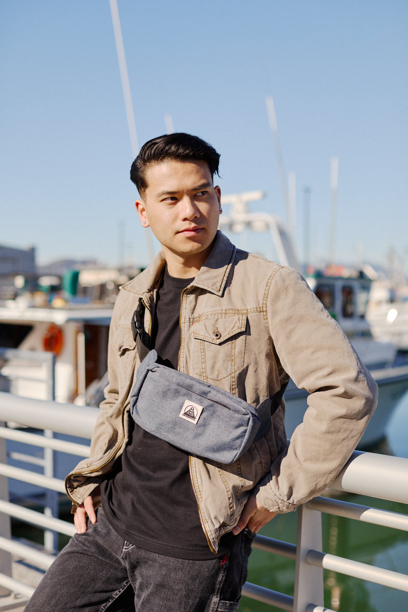 Wearing a tan jacket and black shirt, a person stands by a railing near boats with the Sling Bag, featuring an adjustable strap, casually draped across their chest.