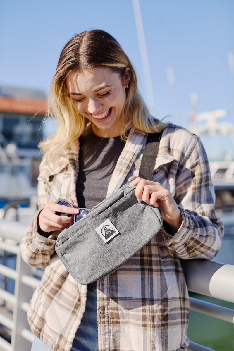 A person smiles while holding the Sling Bag, featuring a patch and adjustable strap, and stands near a railing with boats in the background.