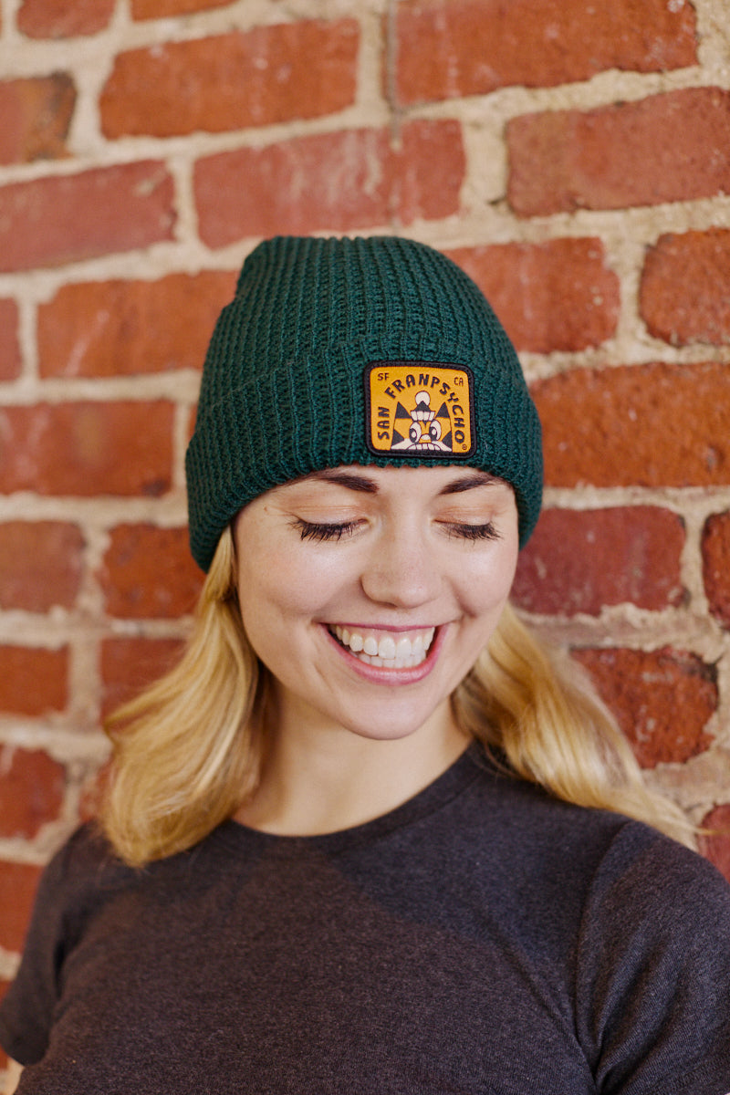 A smiling woman stands against a brick wall, sporting a Cat Stamp Green Waffle Beanie.