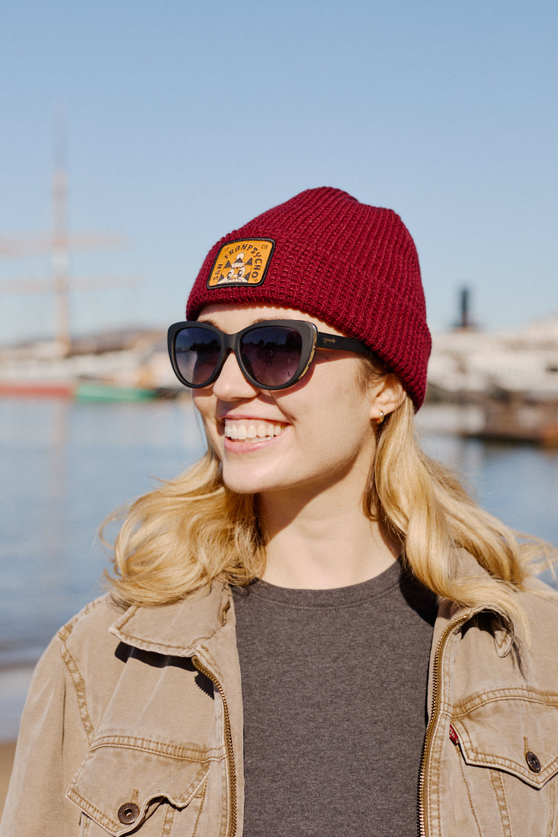 A smiling person in sunglasses and a Cat Stamp Burgundy Waffle Beanie enjoys the waterfront on a clear day.