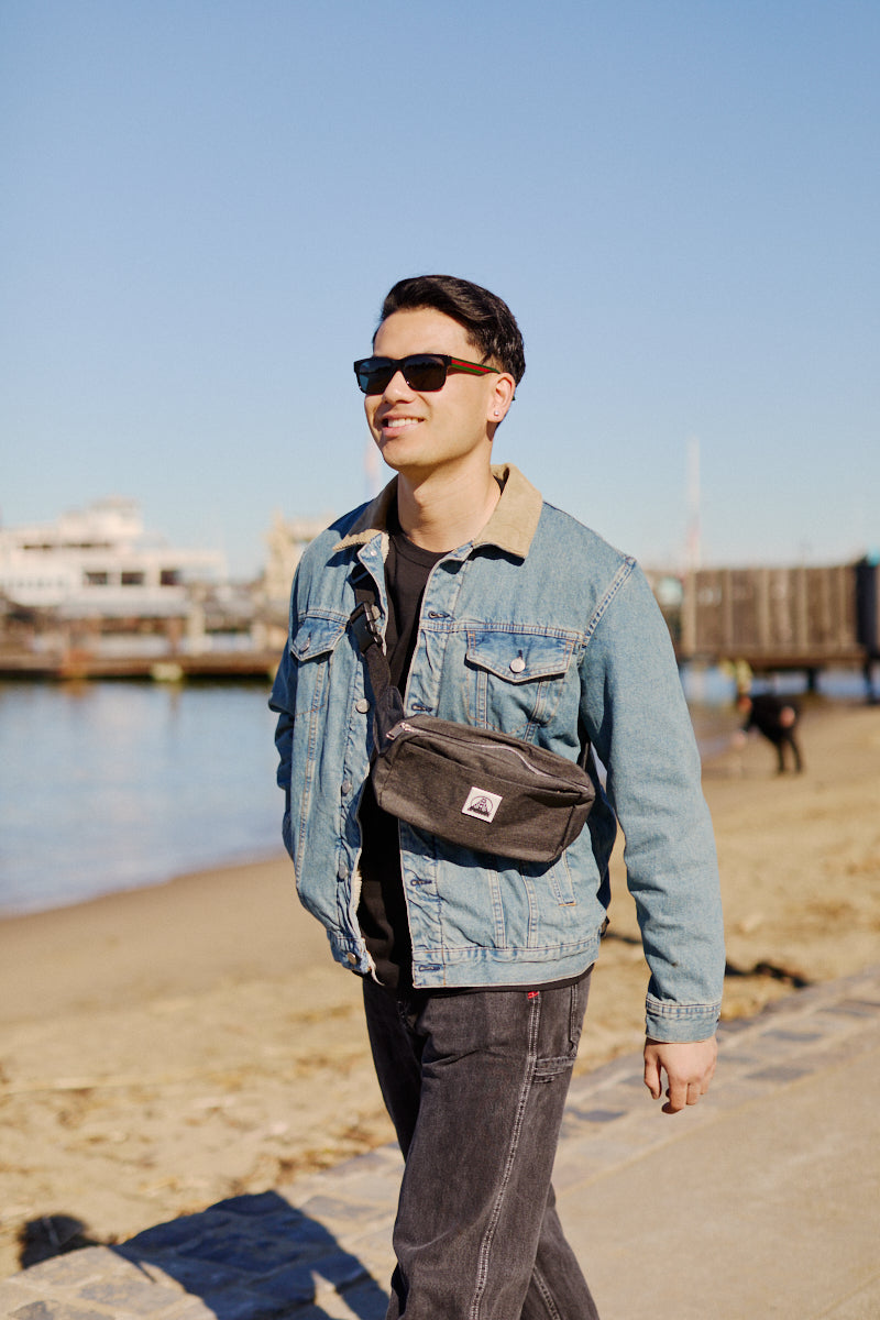 A man in a denim jacket and sunglasses strolls along the sunny beach, his Sling Bag with an adjustable strap swaying gently by his side.