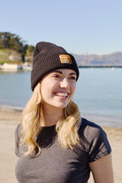 A smiling woman with long hair wears a Cat Stamp Black Waffle Beanie by the waterfront, with a clear sky in the background.