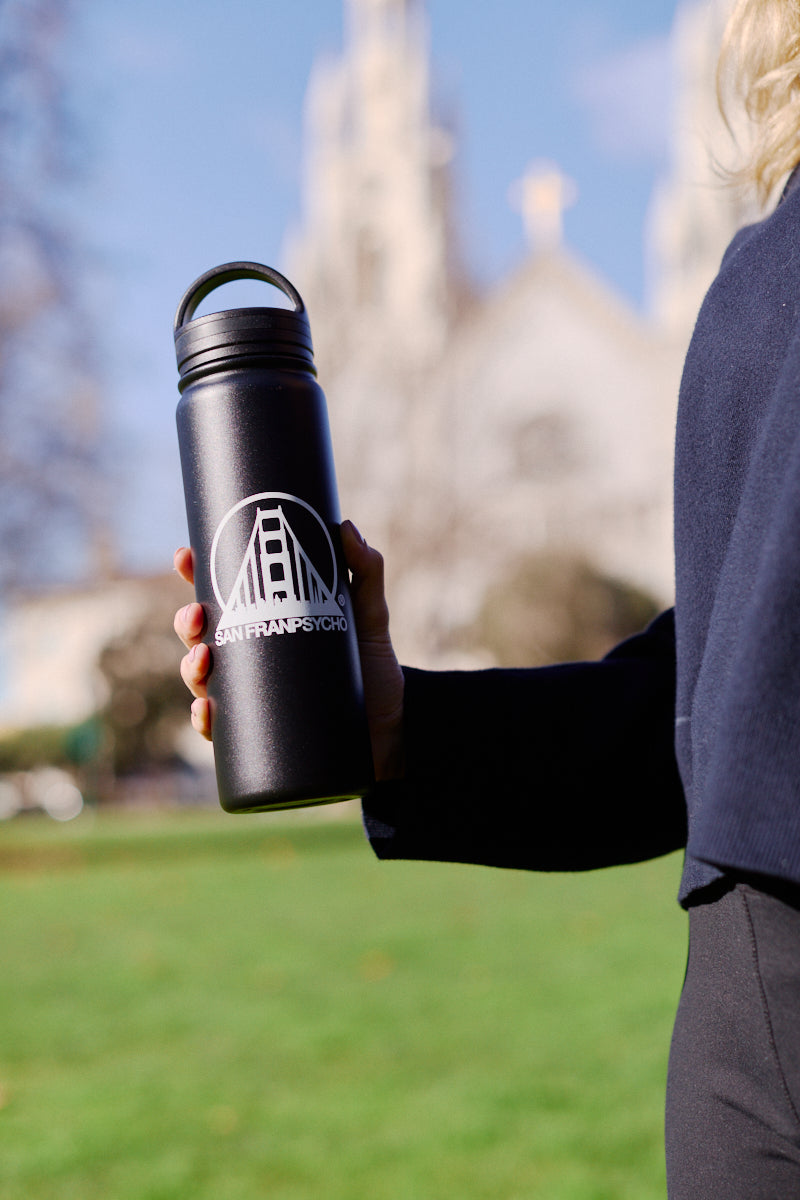 In a grassy area, someone holds the SFP Water Bottle, featuring a sleek stainless steel design with a San Francisco logo. Its double-wall vacuum insulation keeps drinks at the perfect temperature and its leak-resistant feature provides peace of mind. A blurred building looms in the background.