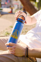 A person outside holds a leak-resistant San Francisco SFP Water Bottle in blue, wearing a white shirt and bracelet.