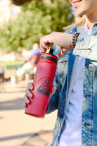 A person in a denim jacket holds the SFP Water Bottle, a stylish red stainless steel bottle featuring a San Francisco bridge design. It offers double-wall vacuum insulation and leak-resistant assurance, making it perfect for outdoor adventures.