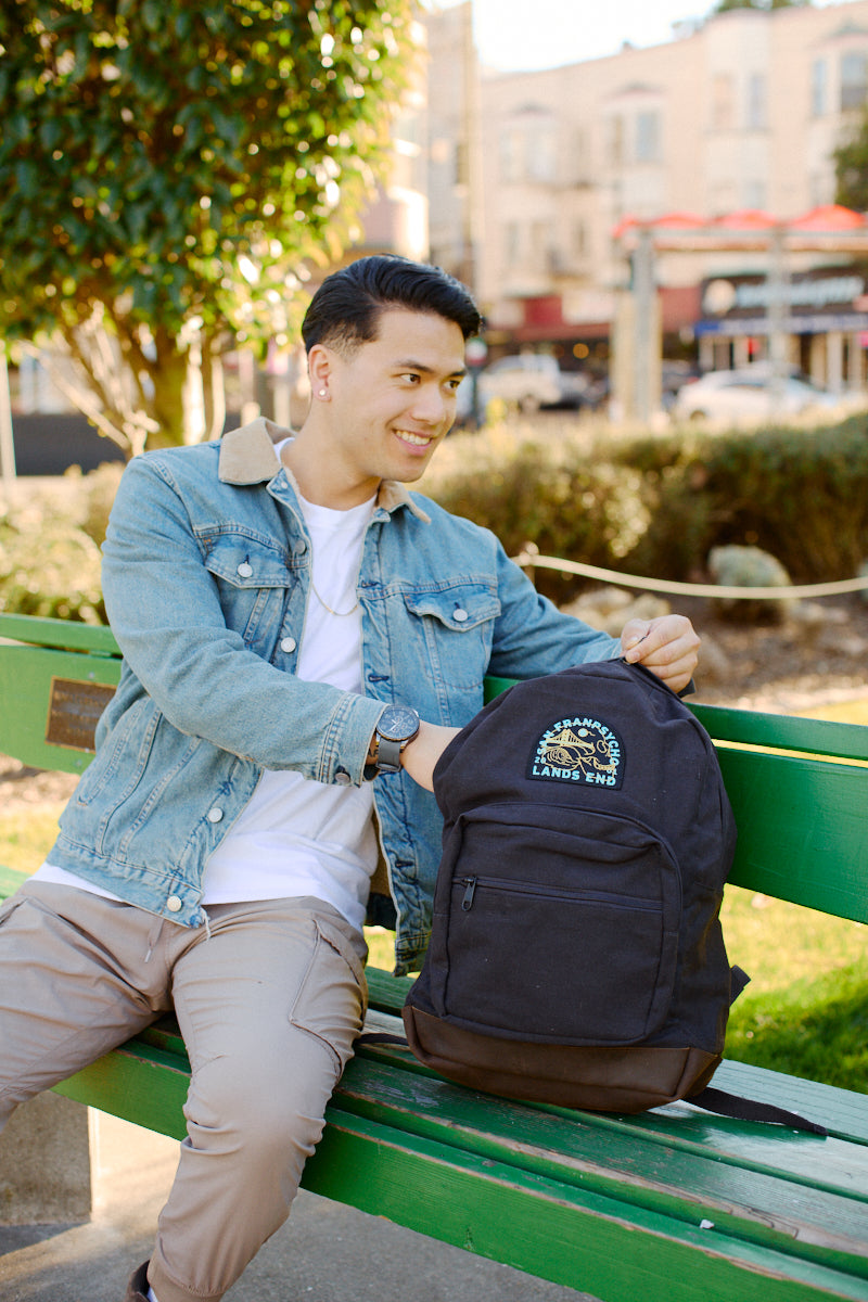 A man in a denim jacket sits on a bench, smiling as he holds the Black Backpack w/ Lands End Patch featuring leather trims.