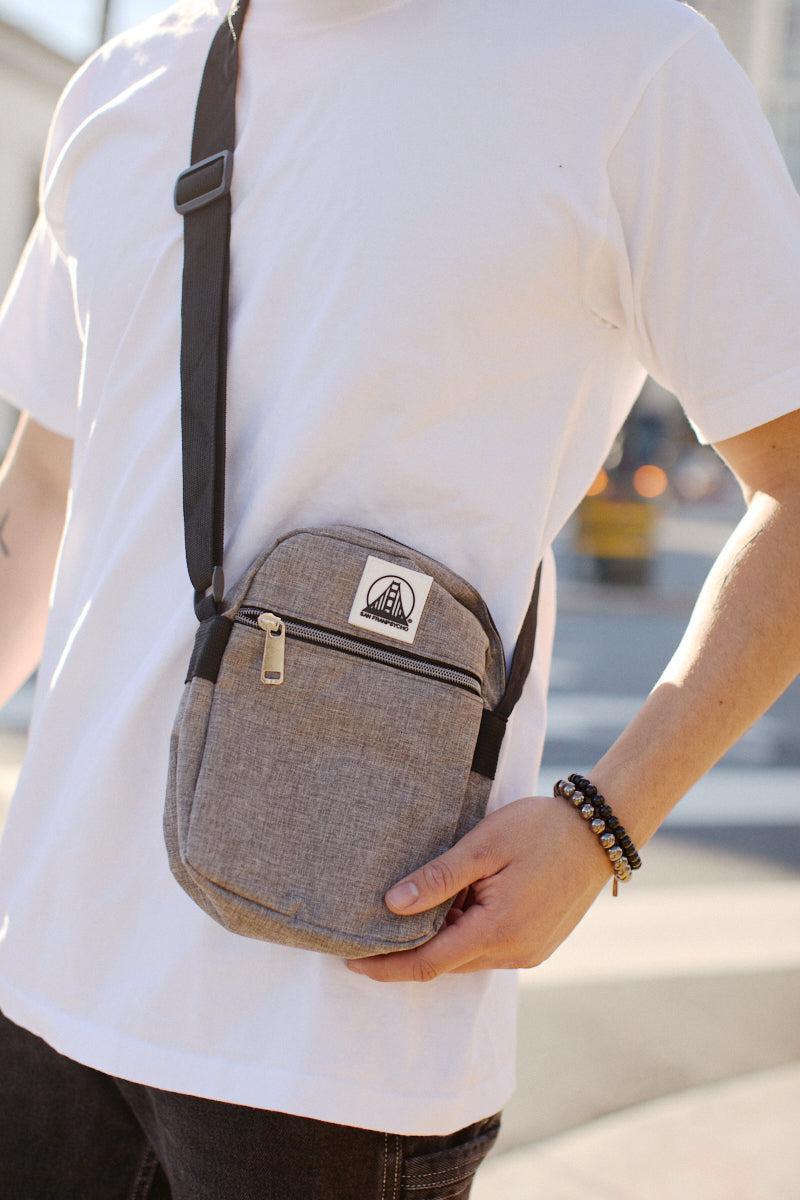 A person wearing a white T-shirt and a Small Sling Bag with an adjustable strap and patch stands outdoors.