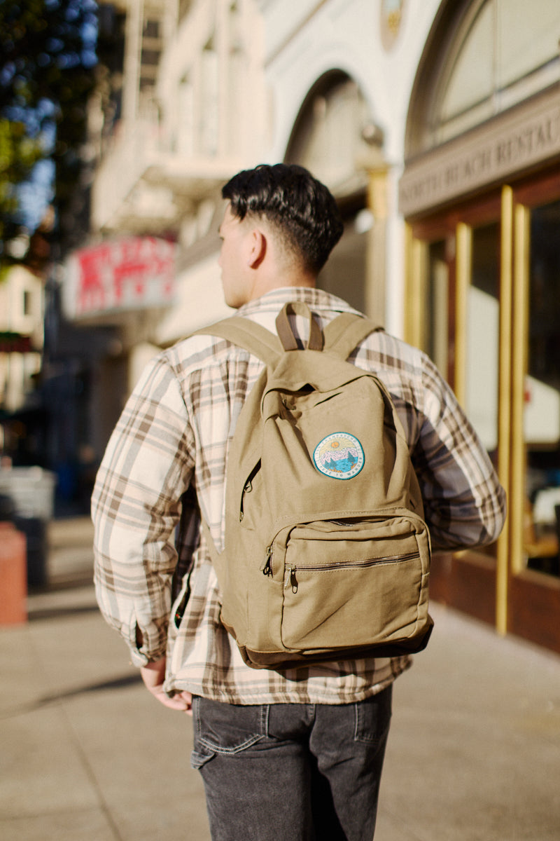 A person with an Olive Backpack w/ Free to Wander Patch strolls down a sunlit sidewalk in the urban area.