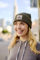 A smiling person in a gray hoodie and Olive Waffle Beanie w/Natural/ Black Logo stands outdoors with a blurred cityscape backdrop.