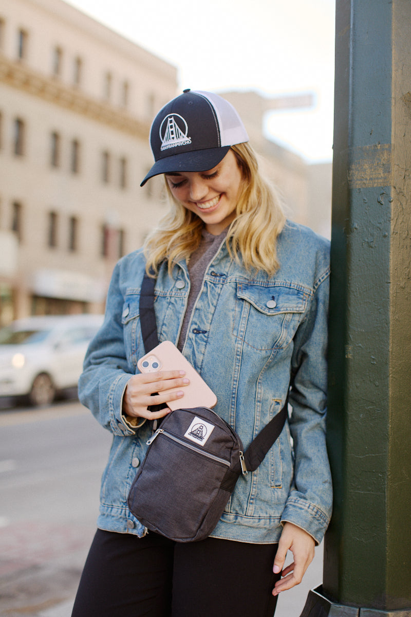 Smiling on a city street, a person in a denim jacket and cap effortlessly slings the Small Sling Bag, featuring an adjustable strap, over one shoulder, combining style and functionality to carry everyday items with ease.