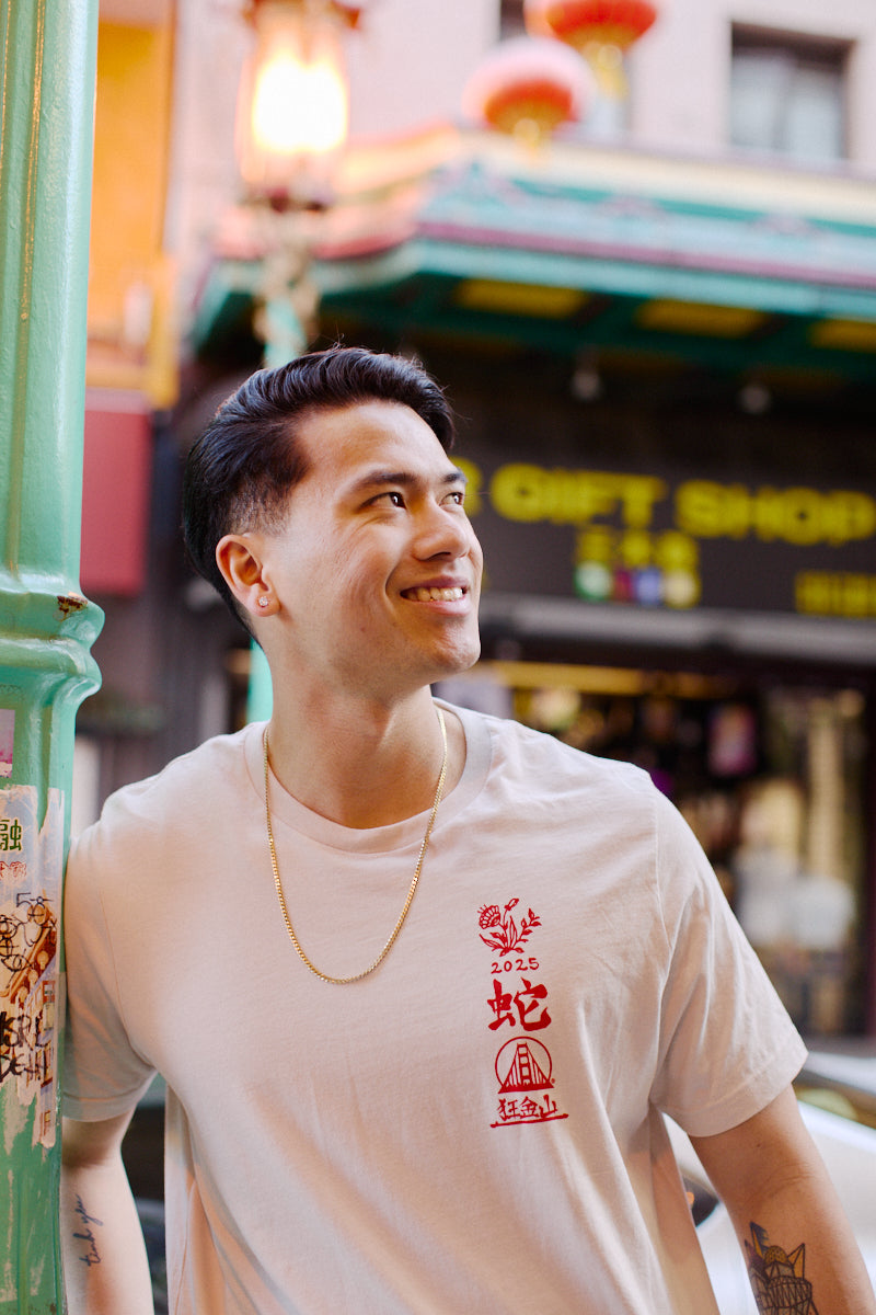 A smiling person wearing the Year of the Snake Tee, designed in a unisex fit and white color, stands by a green pole with a colorful shop facade in the background.