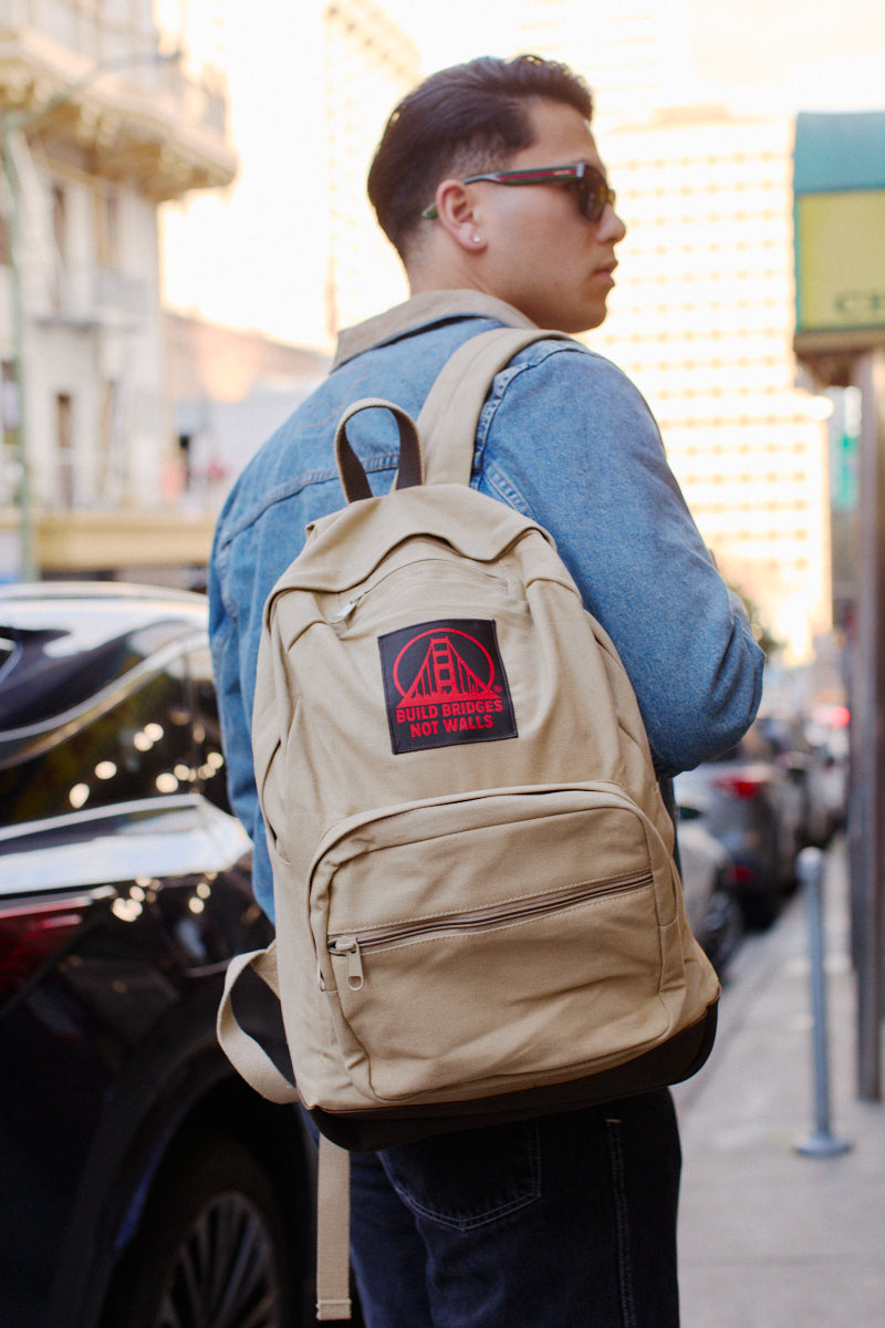 Person wearing sunglasses and a denim jacket, carrying the Khaki Backpack w/ Build Bridges Not Walls Patch featuring leather trims, standing on a city street.