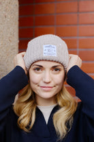 A person, donning an Alabaster Lumberjack Beanie w/ Wet Wreckless and dark sweater, holds the hat while standing against a brick wall, radiating San Franpsycho vibes.