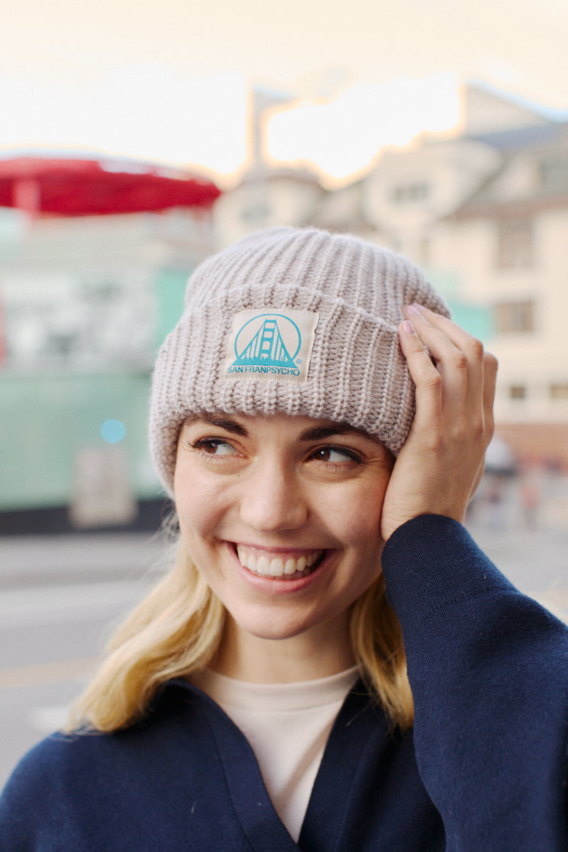 A woman smiles warmly, wearing a dark sweater and an Alabaster Lumberjack Beanie with a Natural/Turquoise Logo, amidst the bustling charm of an urban area.