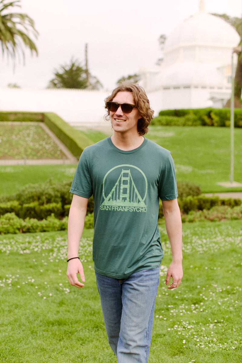 A man in sunglasses, smiling, walks outside on a grassy lawn wearing a limited edition Green Logo Tee.