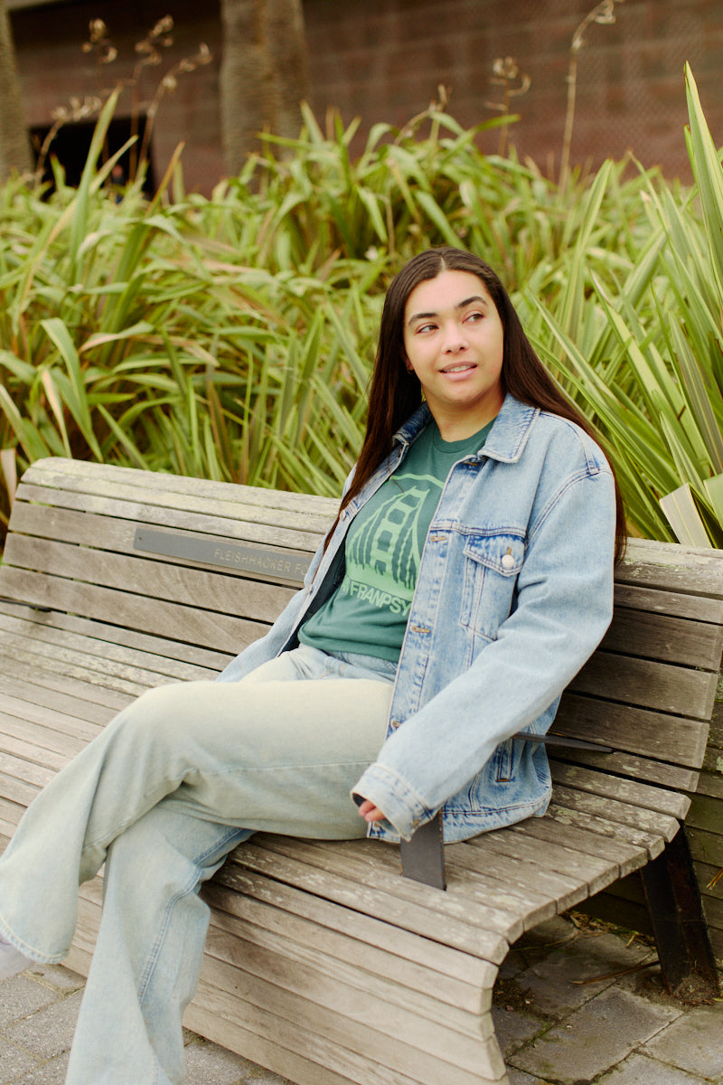 A woman in a Tri-Blend denim jacket and jeans sits on a wooden bench outdoors, surrounded by lush greenery, stylishly showcasing the limited edition Green Logo Tee.