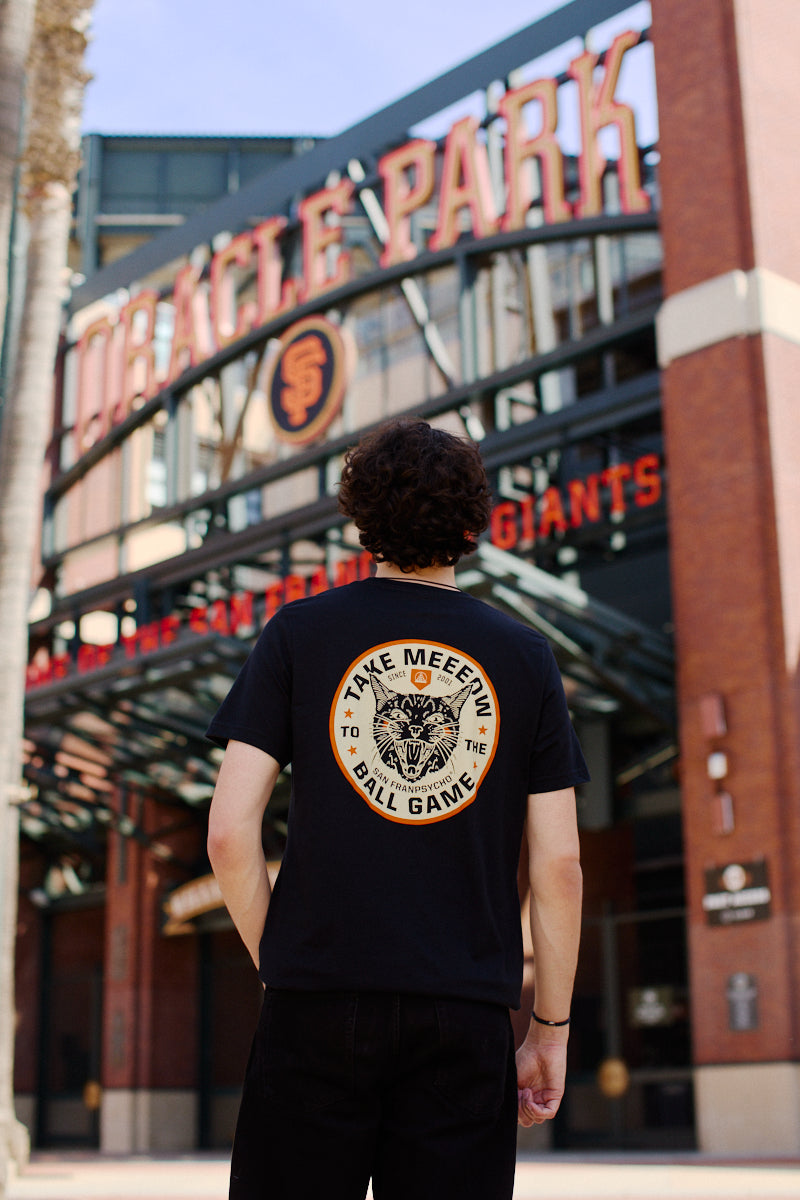 A person in a black Take Meeeow Tee featuring a baseball design stands proudly at the Oracle Park entrance, exuding black and orange pride.