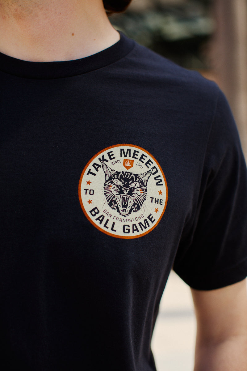 Wearing the Black Take Meeeow Tee, a person showcases a hand-printed circular design featuring a cat and the words Take Meow to the Ball Game, symbolizing black & orange pride.