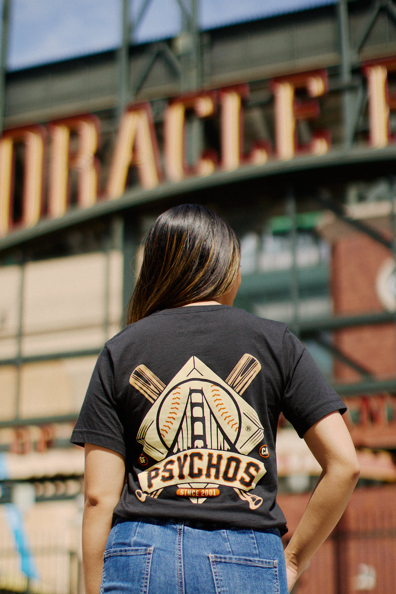 A person in a Psychos Logo Tee, showcasing San Francisco pride with its hand-printed design and black & orange colors, stands by the stadium entrance.